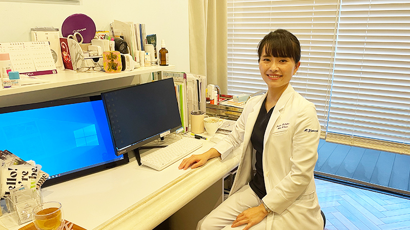 まいこホリスティックスキンクリニックの山﨑まいこ先生が診察室で笑顔で座っている写真。白衣を着た先生の後ろにはデスクがあり、温かみのある診療環境が伝わる一枚。