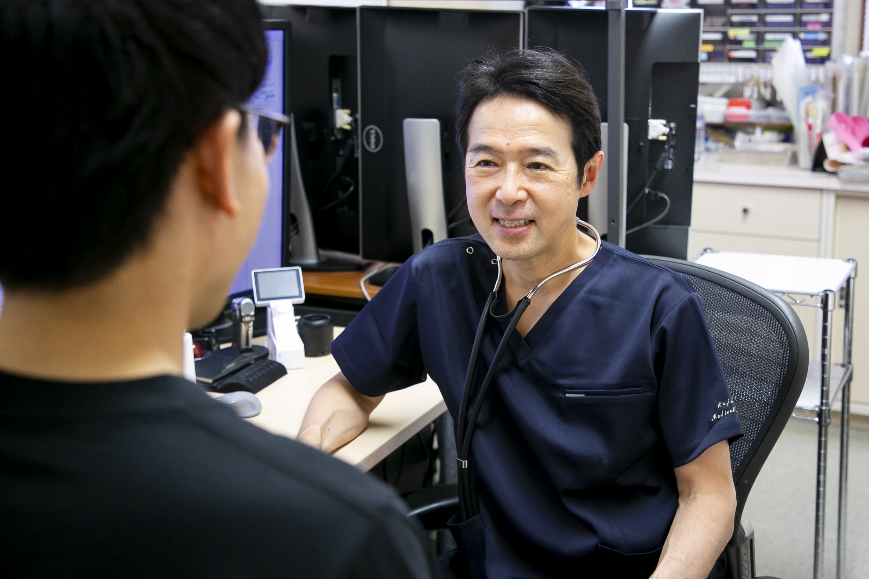 梶の木内科医院・梶尚志先生が診察室で患者と向き合っている様子。笑顔で話しかける表情から、信頼感と親しみが伝わる。